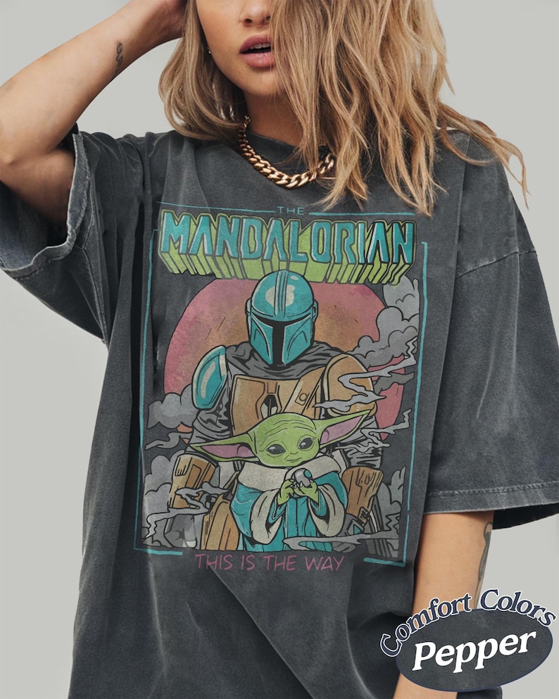 May include: A charcoal gray oversized t-shirt featuring a graphic print of The Mandalorian and Grogu. The design includes the text THE MANDALORIAN at the top and THIS IS THE WAY at the bottom, set against a pink circular background with smoke elements.