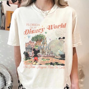 May include: Cream-colored t-shirt featuring a colorful graphic of Disney World in Florida. The design includes Mickey Mouse, a castle, and the text "Florida Disney World Est. 1971" and "The Most Magical Place On Earth."