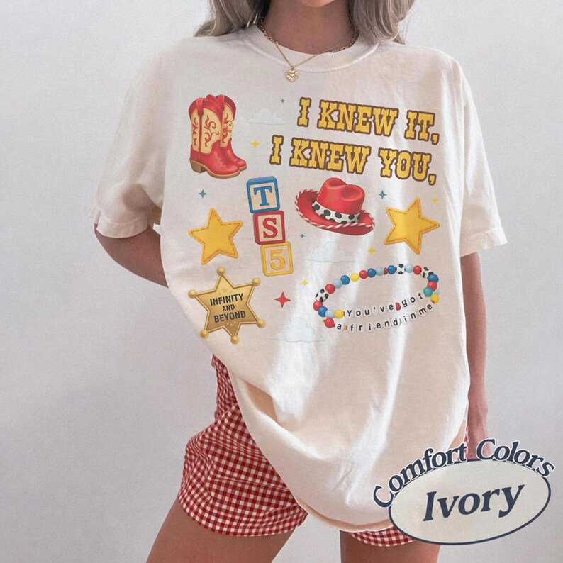 May include: An ivory t-shirt featuring a graphic print with the text I KNEW IT, I KNEW YOU, INFINITY AND BEYOND, and YOU'VE GOT A FRIEND IN ME. The design includes a red cowboy boot, a red cowboy hat, yellow stars, alphabet blocks, and a colorful bead bracelet.