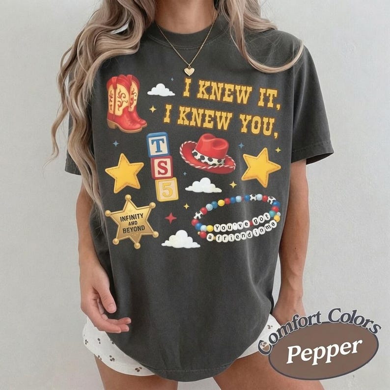 May include: A charcoal gray short-sleeve t-shirt featuring a graphic print with the text I KNEW IT, I KNEW YOU, a red cowboy boot, a red cowboy hat, yellow stars, a sheriff badge with INFINITY AND BEYOND, alphabet blocks, and a beaded bracelet.