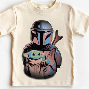K&ouml;nnte beinhalten: Ein cremefarbenes Kurzarm-T-Shirt mit einem Grafikdruck eines Charakters, der einen metallisch blauen und grauen Helm sowie eine R&uuml;stung tr&auml;gt und hinter einer kleinen gr&uuml;nen Kreatur mit gro&szlig;en Ohren und Augen in einer beigen Robe steht.