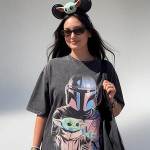 May include: A dark gray t-shirt featuring a graphic of The Mandalorian and Grogu. The shirt is worn with a Grogu-themed headband and sunglasses. A black tote bag is carried over the shoulder. The overall theme is Star Wars.