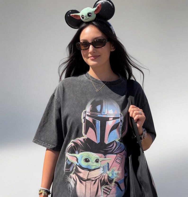 May include: A dark gray t-shirt featuring a graphic of The Mandalorian and Grogu. The shirt is worn with a Grogu-themed headband and sunglasses. A black tote bag is carried over the shoulder. The overall theme is Star Wars.