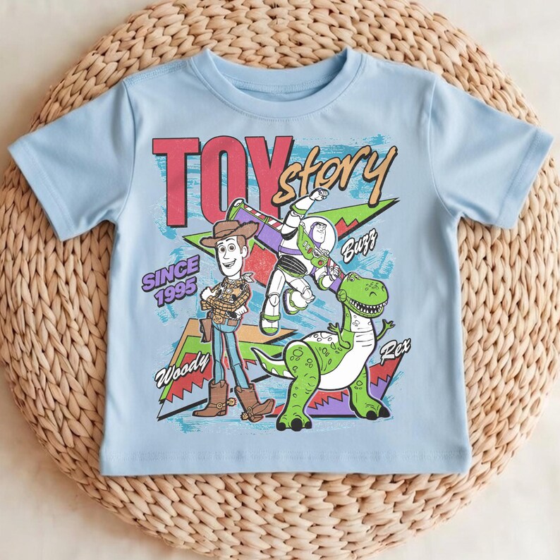 May include: A light blue t-shirt featuring characters from the movie Toy Story. The graphic includes Woody, Buzz Lightyear, and Rex, with the words "TOY STORY" and "SINCE 1995". The shirt is made of a soft material.