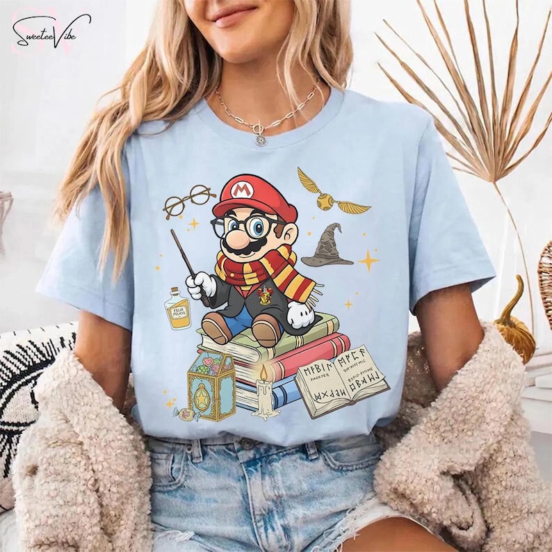 May include: Light blue t-shirt featuring a cartoon Mario character dressed as a wizard, complete with glasses, a wand, and a scarf. The design includes a golden snitch, a wizard hat, and books. The shirt has the text "Sweetee Vibe" at the top.