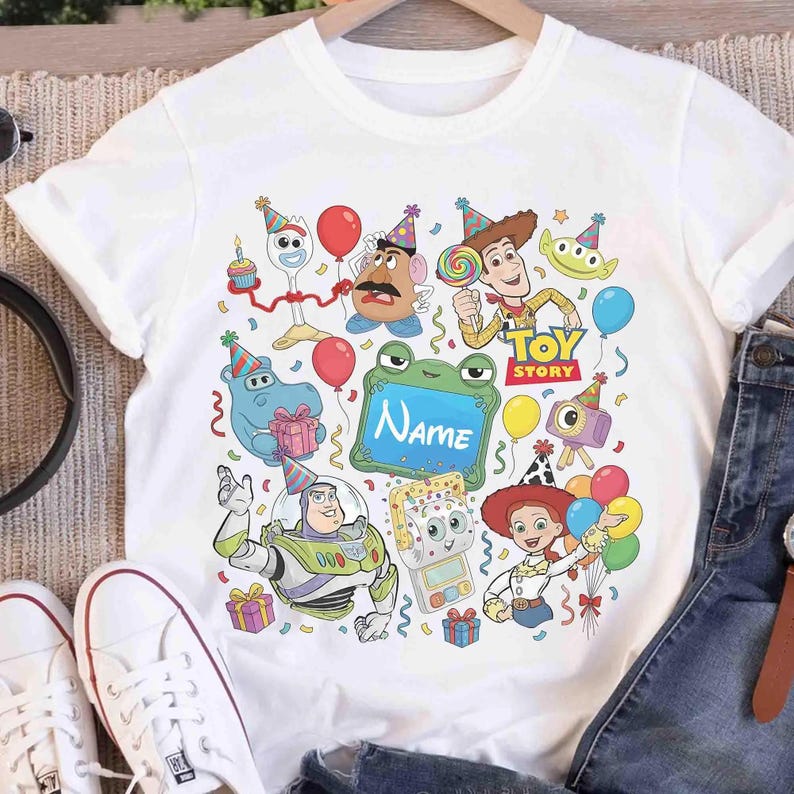 May include: A white short-sleeved t-shirt featuring a colorful graphic print of various Toy Story characters wearing party hats, including Woody, Buzz Lightyear, Jessie, and Mr. Potato Head, with a central green frame containing the text Name.