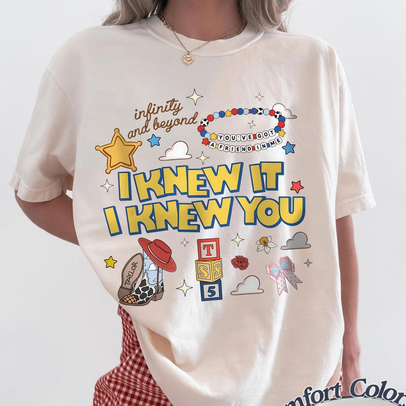 May include: A cream-colored short-sleeve t-shirt featuring a graphic print with the text "infinity and beyond," "YOU'VE GOT A FRIEND IN ME," and "I KNEW IT I KNEW YOU." The design includes a sheriff star, a beaded bracelet, a cowboy boot, and blocks.