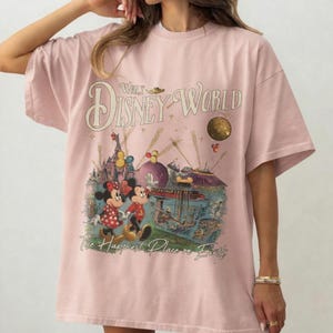 May include: Light pink t-shirt featuring a vintage-style Walt Disney World graphic. The design includes Mickey and Minnie Mouse, a castle, and the phrase "The Happiest Place on Earth."