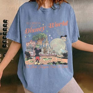 May include: A blue t-shirt featuring a colorful graphic of Disney World in Florida. The design includes the text "Florida Disney World" and images of iconic park landmarks, including Cinderella's castle and Spaceship Earth. Mickey Mouse is also featured.
