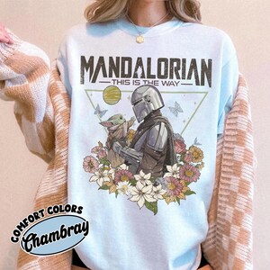 May include: Light blue t-shirt featuring a graphic of The Mandalorian and Grogu surrounded by flowers and butterflies. The text "MANDALORIAN - THIS IS THE WAY" is printed across the top. The shirt has the words "COMFORT COLORS Chambray" printed on the bottom left.