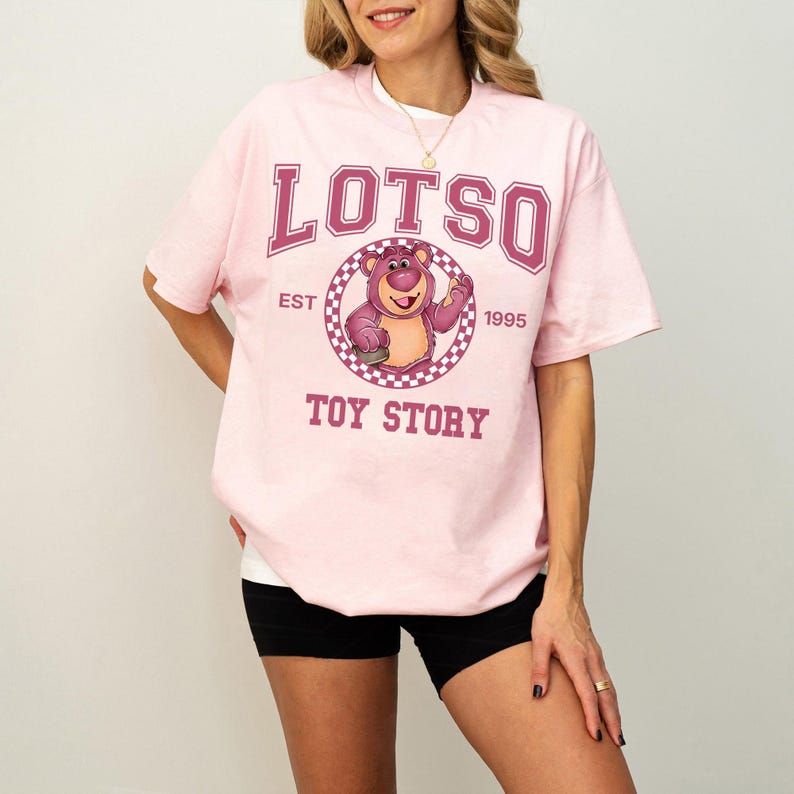 Pu&ograve; includere: T-shirt rosa chiaro con la scritta "LOTSO" in lettere bordeaux sopra una grafica di un orso dei cartoni animati. La grafica &egrave; all'interno di un cerchio a scacchi con "EST 1995" e "TOY STORY" sotto. La maglietta &egrave; girocollo.
