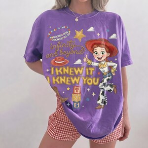 Może przedstawiać: Fioletowy t-shirt z grafiką przedstawiającą kowbojkę z rudymi włosami w kapeluszu oraz napisy "You've got a friend in me", "infinity and beyond" i "I KNEW IT I KNEW YOU", wraz z ilustracjami czerwonego kapelusza, gwiazdy i klock&oacute;w z literami.