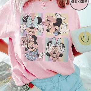 May include: Pale pink crew neck t-shirt with four panels of Minnie Mouse illustrations. Each panel has a different pose and background colour. The shirt has the word "BLOSSOM" in a circle on the upper right. A book and a mug with a smiley face are also visible.