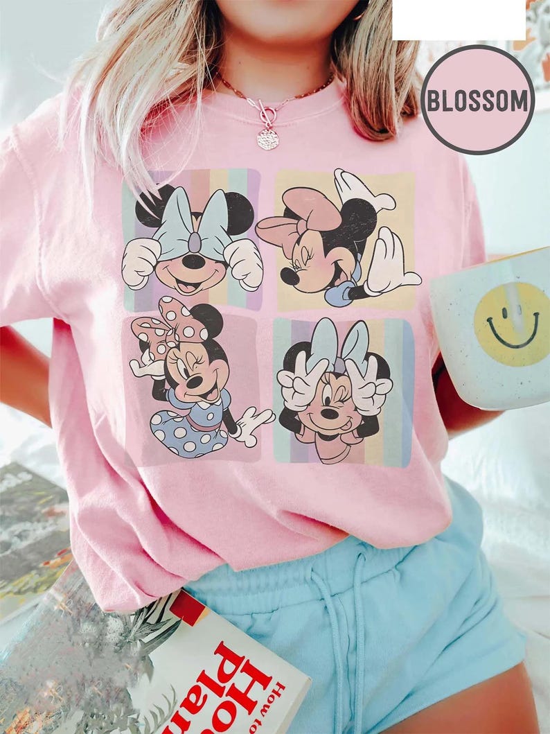 May include: Pale pink crew neck t-shirt with four panels of Minnie Mouse illustrations. Each panel has a different pose and background colour. The shirt has the word "BLOSSOM" in a circle on the upper right. A book and a mug with a smiley face are also visible.