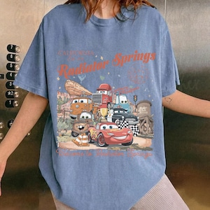 K&ouml;nnte beinhalten: Blaues T-Shirt mit einer Grafik von Charakteren aus dem Film Cars, darunter Lightning McQueen und Mater, mit dem Text "Radiator Springs" in Orange. Das Design umfasst eine W&uuml;stenlandschaft und eine Zielflagge.