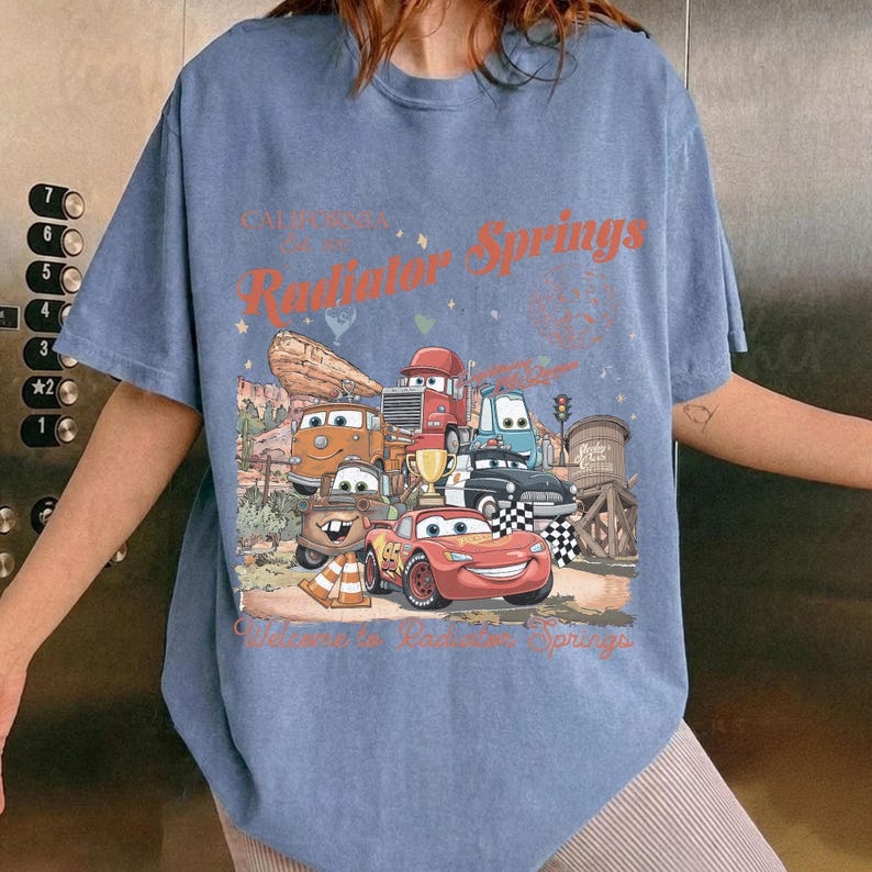 K&ouml;nnte beinhalten: Blaues T-Shirt mit einer Grafik von Charakteren aus dem Film Cars, darunter Lightning McQueen und Mater, mit dem Text "Radiator Springs" in Orange. Das Design umfasst eine W&uuml;stenlandschaft und eine Zielflagge.