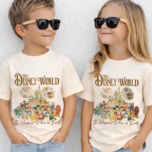 Op de afbeelding: Twee kinderen dragen cr&egrave;mekleurige t-shirts met een Walt Disney World-afbeelding. De afbeelding toont een kasteel, verschillende Disney-personages en de tekst "The Happiest Place on Earth."