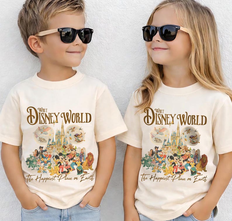 Op de afbeelding: Twee kinderen dragen cr&egrave;mekleurige t-shirts met een Walt Disney World-afbeelding. De afbeelding toont een kasteel, verschillende Disney-personages en de tekst "The Happiest Place on Earth."