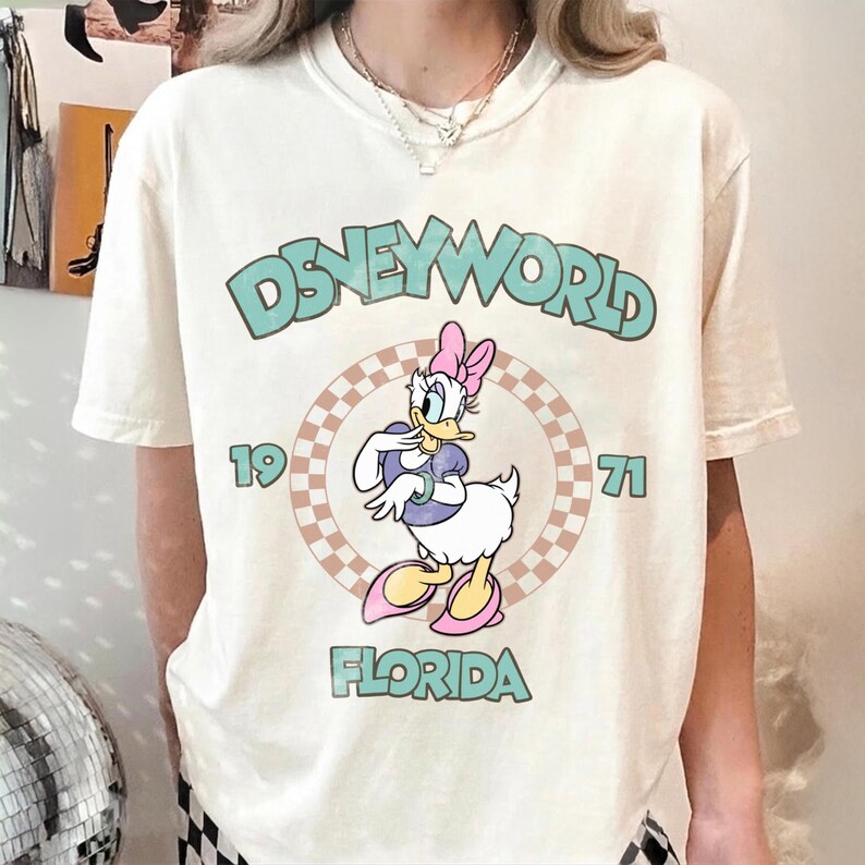 May include: Cream-colored t-shirt featuring a Disney World graphic. The design includes the text "DISNEYWORLD" in teal, the year "1971", and "FLORIDA" in teal. A cartoon of Daisy Duck is in the center, wearing a pink bow and shoes.