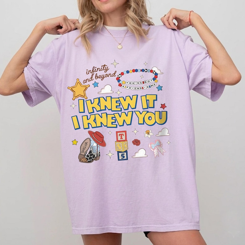 May include: A light purple t-shirt featuring a graphic print with the text "infinity and beyond", "I KNEW IT I KNEW YOU", and "YOU'VE GOT A FRIEND IN ME". The design includes a beaded bracelet, a cowboy boot, alphabet blocks, stars, and clouds.