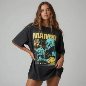 以下が含まれることがあります： マンダロリアンとグローグのグラフィックが特徴の黒いTシャツ。「MANDO」の文字が黄色のブロック体で、フレーズ「THE LEGEND CONTINUES」がデザインされています。このシャツは半袖で、ゆったりとしたフィット感です。