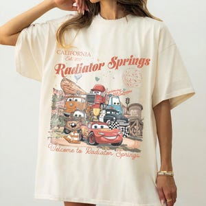 K&ouml;nnte beinhalten: Cremefarbenes T-Shirt mit einer Grafik von Charakteren aus dem Film Cars, mit dem Text "Radiator Springs" und "Welcome to Radiator Springs". Das Design beinhaltet eine W&uuml;stenlandschaft und die Worte "California Est. 2012".