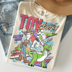 May include: Cream-colored t-shirt featuring the words "Toy Story" in pink and red, with cartoon characters Woody, Buzz, and Rex. The shirt also has the text "Since 1995". The shirt is folded on top of blue jeans.
