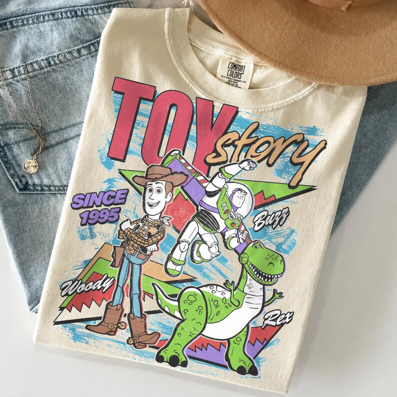 May include: Cream-colored t-shirt featuring the words "Toy Story" in pink and red, with cartoon characters Woody, Buzz, and Rex. The shirt also has the text "Since 1995". The shirt is folded on top of blue jeans.