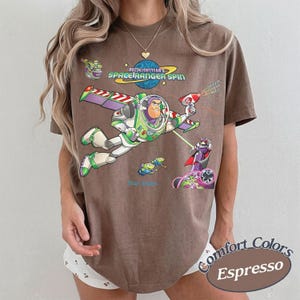 May include: A brown short-sleeve t-shirt featuring a graphic print of Buzz Lightyear from Space Ranger Spin. The design includes Buzz Lightyear flying, a small alien, and the text Buzz Lightyear's Space Ranger Spin. The shirt has a crew neckline.
