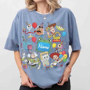 May include: A dusty blue t-shirt featuring a colorful Toy Story graphic print. The design includes characters like Woody, Buzz Lightyear, Jessie, and Forky wearing party hats, surrounded by balloons, gifts, and a central frame with the text Name.