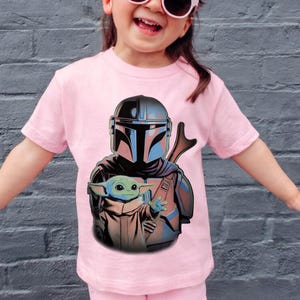K&ouml;nnte beinhalten: Ein hellrosa T-Shirt mit kurzen &Auml;rmeln und einem Grafikdruck, der einen Mandalorianer mit metallischem Helm zeigt, der hinter einer kleinen gr&uuml;nen Kreatur mit gro&szlig;en Ohren und Augen steht, dargestellt in einer farbenfrohen Illustration.