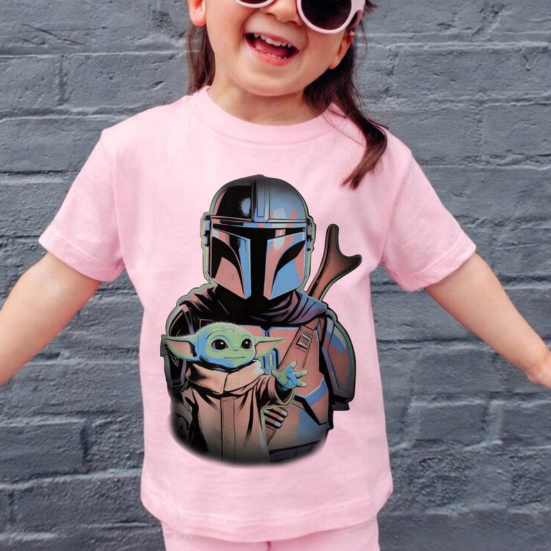 K&ouml;nnte beinhalten: Ein hellrosa T-Shirt mit kurzen &Auml;rmeln und einem Grafikdruck, der einen Mandalorianer mit metallischem Helm zeigt, der hinter einer kleinen gr&uuml;nen Kreatur mit gro&szlig;en Ohren und Augen steht, dargestellt in einer farbenfrohen Illustration.