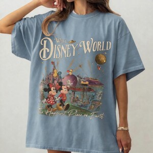 May include: Light blue t-shirt featuring a vintage-style Walt Disney World graphic. The design includes Mickey and Minnie Mouse, a castle, and the phrase "The Happiest Place on Earth."
