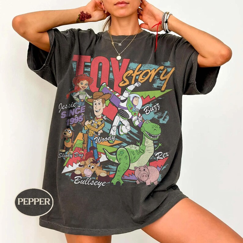 May include: A charcoal gray short-sleeve t-shirt featuring a Toy Story graphic print with characters Jessie, Woody, Buzz Lightyear, Slinky Dog, Bullseye, and Rex. The text includes Toy Story, Jessie, Buzz, Woody, Slinky Dog, Bullseye, and Rex.