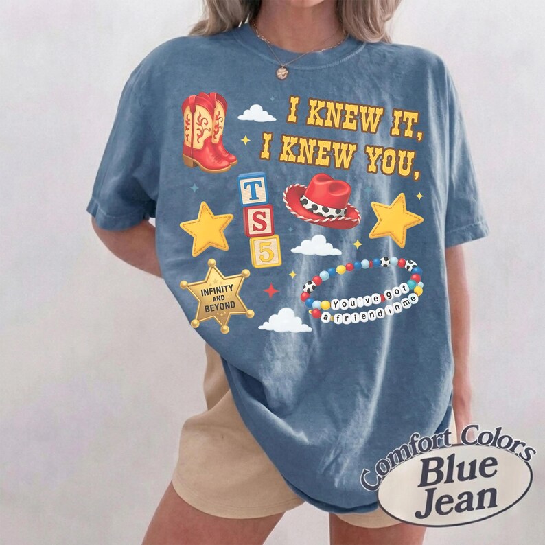 May include: A blue short-sleeve t-shirt featuring a graphic print with the text I KNEW IT, I KNEW YOU, INFINITY AND BEYOND, and YOU'VE GOT A FRIEND IN ME. The design includes red cowboy boots, a red cowboy hat, yellow stars, blocks, and beads.