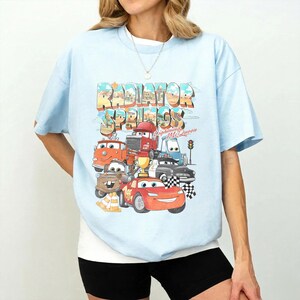 May include: Light blue t-shirt featuring a colorful graphic of characters from the movie Cars, with the text "Radiator Springs" in a retro design. The design includes Lightning McQueen and other vehicles.