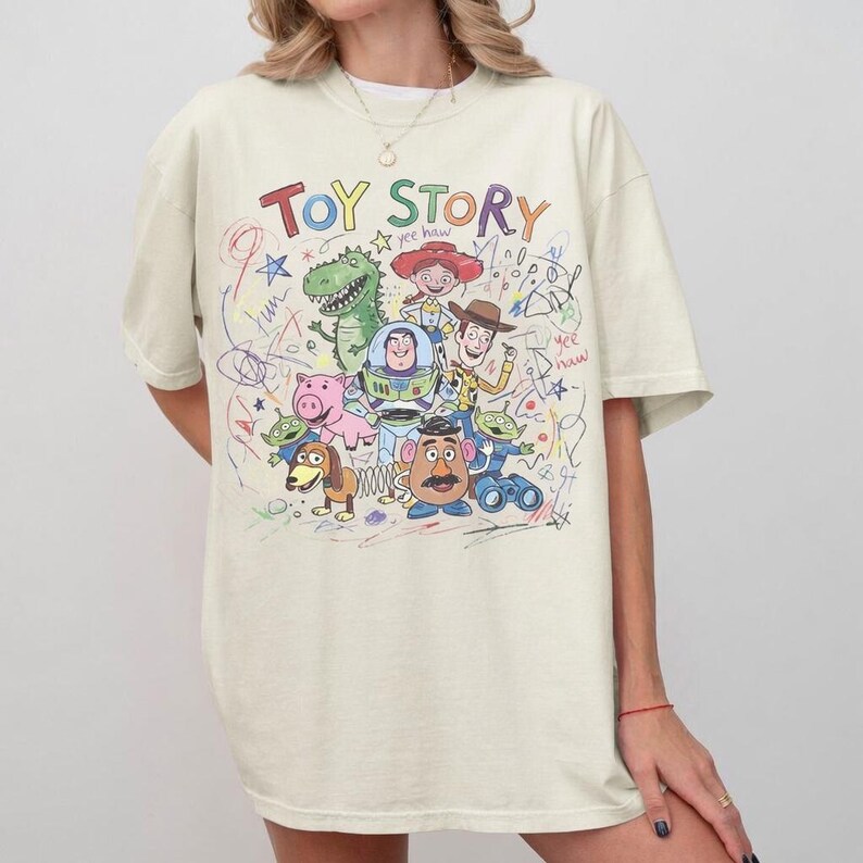May include: Cream-colored t-shirt featuring the words "Toy Story" in rainbow colors, with a colorful cartoon graphic of characters from the movie. The design includes a dinosaur, Buzz Lightyear, Woody, and other characters.