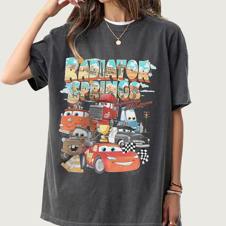 May include: Dark gray t-shirt featuring a colorful graphic of characters from the movie Cars, including Lightning McQueen. The text "Radiator Springs" is displayed above the characters.