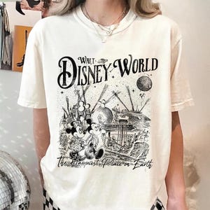 May include: Cream-colored t-shirt featuring a black and white graphic of a vintage Walt Disney World design. The design includes Mickey and Minnie Mouse, a castle, and the text "Walt Disney World" and "The Happiest Place on Earth."