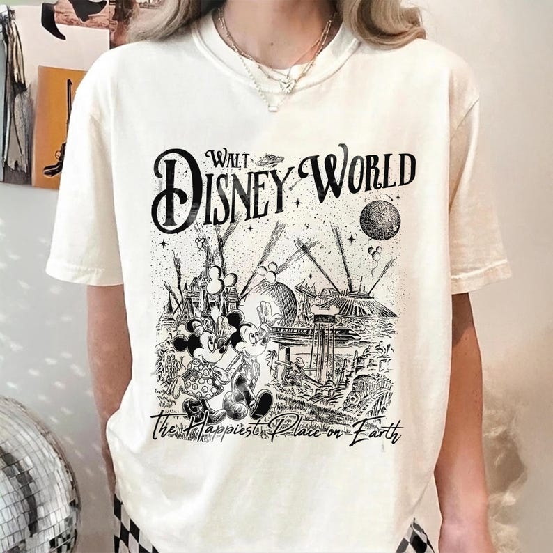 May include: Cream-colored t-shirt featuring a black and white graphic of a vintage Walt Disney World design. The design includes Mickey and Minnie Mouse, a castle, and the text "Walt Disney World" and "The Happiest Place on Earth."