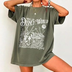 May include: Olive green t-shirt featuring a graphic of Mickey and Minnie Mouse at Walt Disney World. The design includes the words "Walt Disney World" and "The Happiest Place on Earth" in white lettering. The shirt has short sleeves and a relaxed fit.