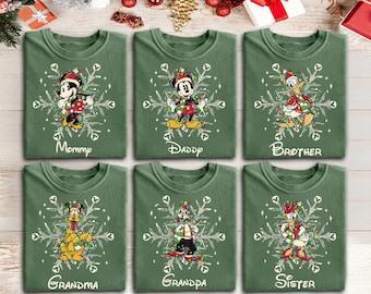 May include: Six olive green crewneck sweatshirts featuring Disney characters wrapped in colorful Christmas lights over a white snowflake design. Each shirt displays a character name: Mommy, Daddy, Brother, Grandma, Grandpa, and Sister.