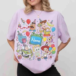 May include: A light purple t-shirt featuring a colorful Toy Story graphic with characters like Woody, Buzz Lightyear, Jessie, and Mr. Potato Head wearing party hats. The design includes balloons, gifts, and a central green sign with the text Name.