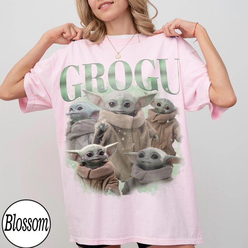 May include: A light pink t-shirt featuring the word "GROGU" in green, with multiple Grogu character illustrations. The design includes a light green watercolor effect. The shirt is a casual, oversized style.
