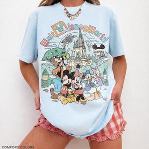 May include: Light blue t-shirt with a colorful Walt Disney World graphic featuring Mickey Mouse, Minnie Mouse, Donald Duck, and other characters. The shirt has the text "Walt Disney World" in a retro font. The bottom of the shirt has the text "CHAMBRAY".