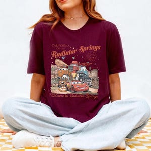 K&ouml;nnte beinhalten: Eine wei&szlig;e Keramiktasse mit schwarzem Henkel und dem Text "But First, Coffee" in fetter, schwarzer Schrift. Die Tasse steht vor einem verschwommenen Hintergrund, der eine gem&uuml;tliche Umgebung suggeriert.