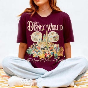 May include: Burgundy t-shirt featuring a colorful graphic of Disney characters and the text "Walt Disney World" and "The Happiest Place on Earth". The graphic includes Mickey Mouse, Donald Duck, and other beloved characters.