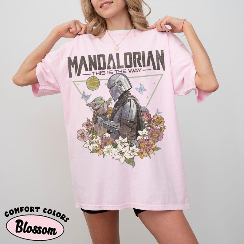May include: A light pink t-shirt featuring a graphic of The Mandalorian and Grogu surrounded by flowers and butterflies. The text "MANDALORIAN - THIS IS THE WAY" is printed across the top. The shirt has the words "COMFORT COLORS" and "Blossom" printed on the bottom.