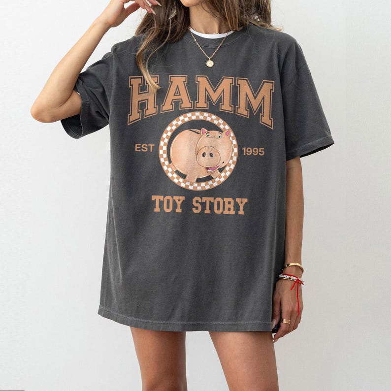 Pu&ograve; includere: T-shirt grigio scuro con la scritta "HAMM" in lettere marroni a blocchi sopra una grafica circolare di un maiale dei cartoni animati. La grafica &egrave; circondata da un bordo a scacchi, con "EST 1995" e "TOY STORY" stampati sotto.
