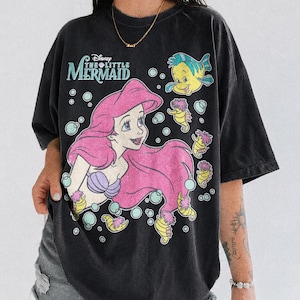 May include: A black short-sleeve t-shirt featuring a graphic print of Ariel from The Little Mermaid with pink hair, a purple seashell top, and Flounder. The design includes bubbles and small seahorses, with the text Disney The Little Mermaid.